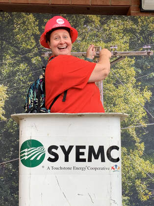 A women in a simulated bucket truck wearing a hard hat. The Surry-Yadkin Electric Membership Corporation logo is on the front of the bucket.