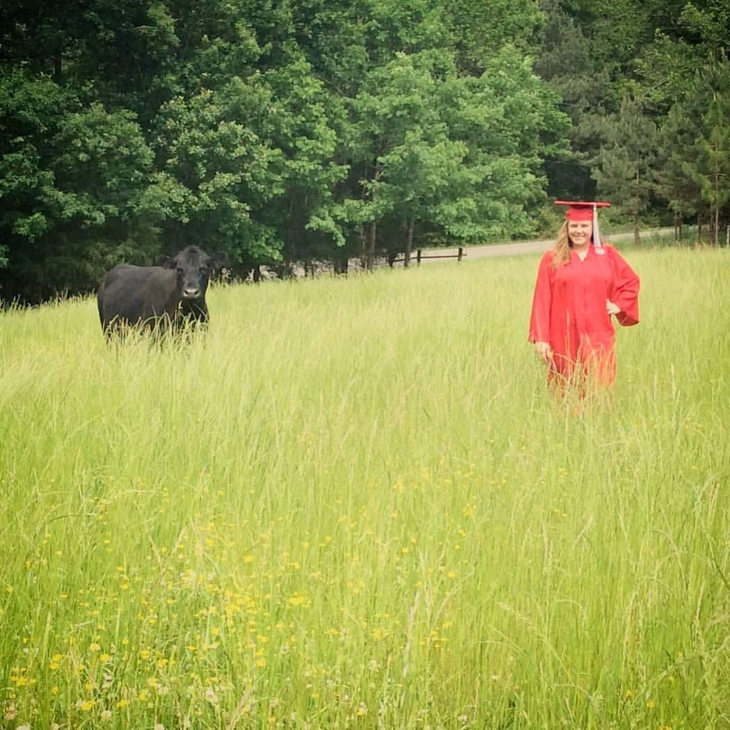 Sable White wearing a graduation cap and gown standing in a field next to a cow.