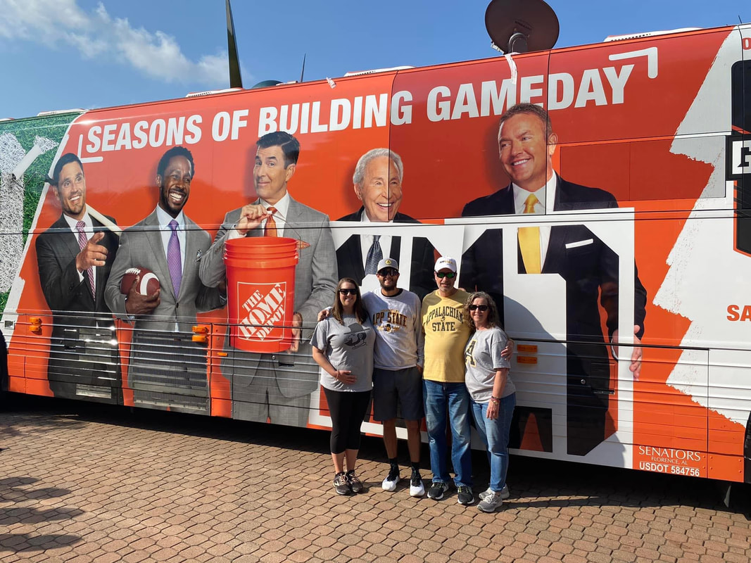 Four people standing together in front of a tour bus. The bus shows an ad for Home Depot. The ad consists of a photo of five men standing side by side. The man in the center is holding a Home Depot bucket. The ad text says: Seasons of building gameday.
